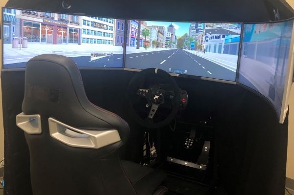 Driving simulator with three curved high-definition monitors in a semi-circle configuration. A racing-car seat with steering wheel and foot pedals in front of monitor setup.